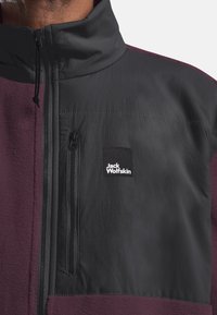 Fleecejacke in Burgunderrot und Schwarz, mit hohem Kragen, Reißverschlusstasche und Logo-Patch auf der Brust. Glatte Textur, strapazierfähiges Design.