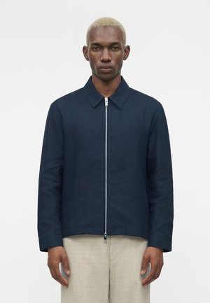 Lühikeste blondide juustega mees kannab tumesinist lukuga jopet kraega ja heledat beežid pükse, seistes ühevärvilise valge tausta ees.