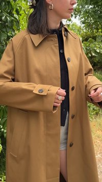 Cappotto leggero marrone con taglio dritto, ampio colletto, chiusura a bottoni e due tasche laterali, indossato sopra un top scuro. Tessuto strutturato.