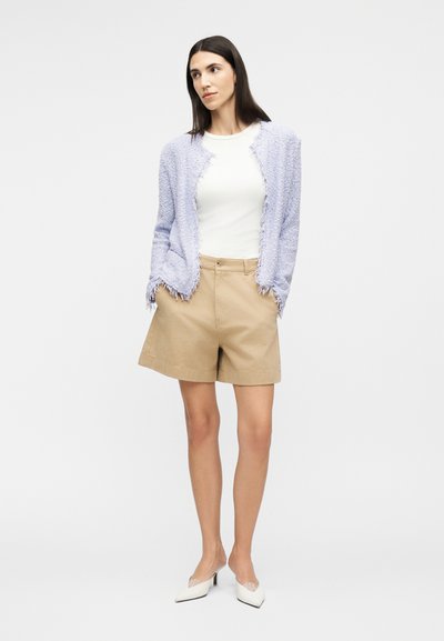 Γυναίκα που στέκεται με τα χέρια στις τσέπες, φοράει λευκή μπλούζα, πλεκτή ανοιχτό μωβ ζακέτα, μπεζ σόρτς και λευκές μυτερές γόβες.