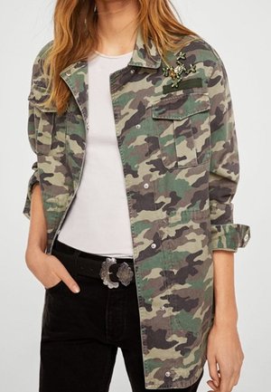 Žena v zelené maskáčové bundě s korálkovou broží ve tvaru ještěrky, v bílém tričku, černých kalhotách a ozdobné stříbrné přezce na opasku, stojící s jednou rukou v kapse.