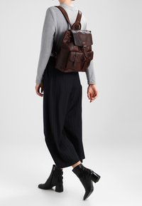 Brun läderryggsäck med justerbara remmar, ett topplock och framfickor. Bärs över kolgrå vida byxor och ankelboots.