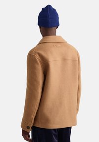 Bruine wolblend jas met een gestructureerde kraag, knoopmanchetten en een achternaad detail. Gecombineerd met een geribbelde blauwe gebreide beanie.