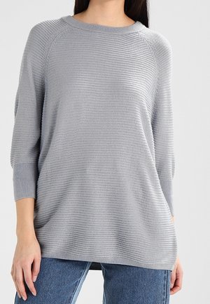 Woman wearing a light gray ribbed knit sweater with 3/4 sleeves and blue jeans against a plain light background.