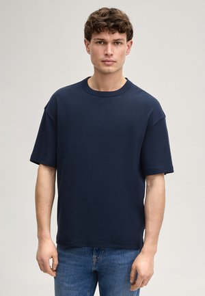 Jeune homme aux cheveux courts et bouclés, portant un t-shirt bleu marine uni à manches courtes et un jean bleu, debout devant un fond neutre.