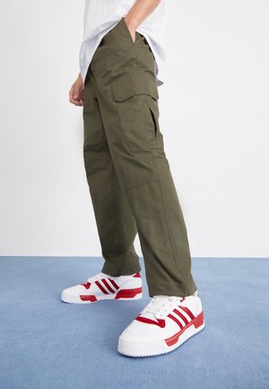Persoon die olijfgroene cargobroek en witte sneakers met rode strepen draagt, staand op een blauw tapijt tegen een witte achtergrond.