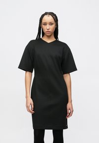 Μαύρο φόρεμα με κοντό μανίκι, V-λαιμόκοψη, εφαρμοστή σιλουέτα, λεία υφή και μινιμαλιστικό σχέδιο, που φτάνει μέχρι τα μέσα των μηρών.