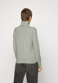 Hellegrauer Rollkragenpullover mit tailliertem Schnitt, langen Ärmeln und einer glatten, weichen Textur, getragen über dunkelgrauen Jeans.