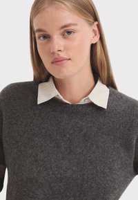 Donkergrijs sweater met een textuurafwerking, met daaronder een witte blouse met een subtiele streepjespatroon.