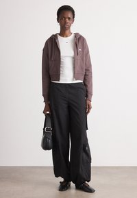 Avskåret brun hettejakke med glidelås, hvit tettsittende skjorte, svarte vidde bukser med cargo-lommer, svarte flate sko, og en svart håndveske.