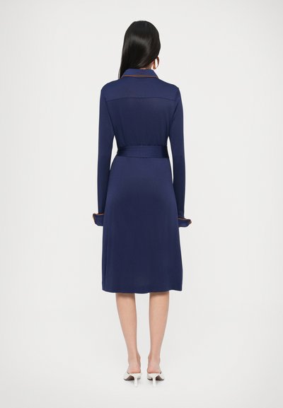 Μπλε φόρεμα ναυτικού με μακριά μανίκια, ζώνη στη μέση και πορτοκαλί λεπτομέρειες στα μανίκια. Λαμπερό ύφασμα με γιακά και μήκος μέχρι το γόνατο. Απλός σχεδιασμός.