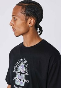 Camiseta negra con un estampado gráfico que presenta colores pastel y texto, mangas cortas y cuello redondo. Peinada con trenzas.