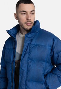 Modrá nafukovacia bunda s vysokým golierom, ktorá má prešité vzory a zipsové zapínanie. Tkanina má jemný lesk a hladkú textúru.