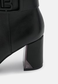 Bota de tobillo de cuero negro con tacón cuadrado y detalle de logo metálico en el lateral del tacón, sobre un fondo blanco.