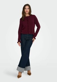 Pull en tricot bordeaux avec un ourlet à volants, détails de boutons, associé à un jean évasé foncé avec un revers contrastant, et des chaussures à bouts pointus marron.