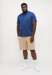 Tricou bleumarin cu mânecă scurtă, cu un logo mic, asortat cu pantaloni scurți bej, teniși albi și șosete albe; croială lejeră.