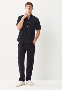 Camicia nera a maniche corte con bottoni e taschino sul petto, abbinata a jeans neri larghi e sneakers bianche su sfondo neutro.