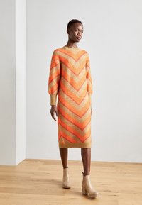 Robe en tricot orange et beige avec un motif en chevrons, dotée de manches ballons et d'une silhouette droite. Portée avec des bottines beiges.