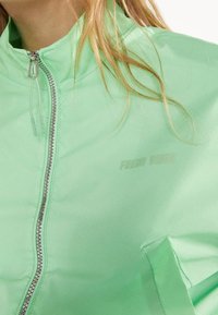 Mintgrön zip-up jacka i mjukt, lätt tyg. Har en hög krage, silverdragkedja och "FRESH VIBES" präglat p å bröstet.