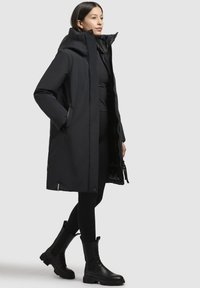 Parka noire isolée avec un col haut, fermeture éclair à l'avant, poches latérales et texture lisse. Accompagnée de leggings noirs et de bottillons.