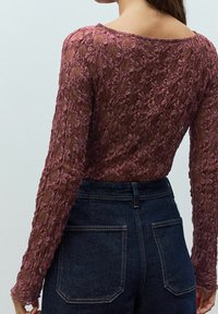 Femme portant un haut à manches longues en dentelle florale rose semi-transparente et un jean en denim bleu foncé taille haute, vue de dos.