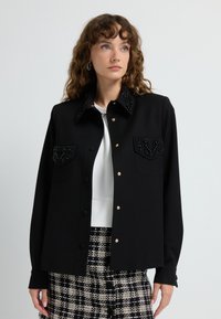 Black jacket with beaded collar and chest pockets, featuring gold buttons and long sleeves. Worn over a white top, paired with patterned skirt.