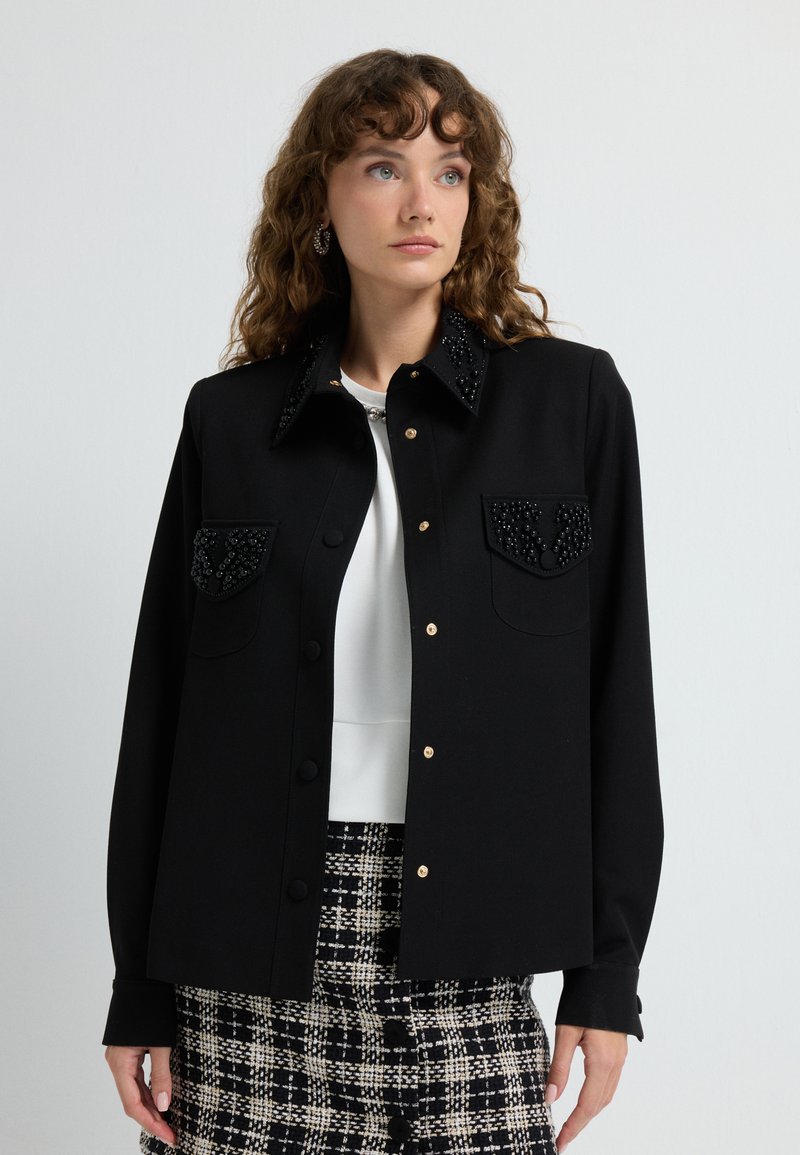 Black jacket with beaded collar and chest pockets, featuring gold buttons and long sleeves. Worn over a white top, paired with patterned skirt.