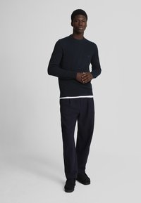 Marinblå stickad tröja med rund hals, tillsammans med svarta byxor. Modellen står med händerna ihopknäppta och visar en jämn textur och passform.