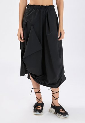 Gonna midi nera asimmetrica con orlo arricciato, indossata con sneakers nere stringate con suole spesse bianche.