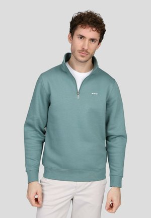Sweatshirt zippé en teal clair fabriqué dans un matériau doux, avec un col haut, des poignets côtelés et un petit logo sur la poitrine.