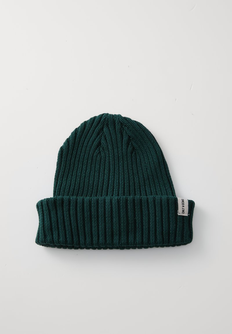 Bonnet en tricot côtelé vert avec un revers. Présente un motif texturé et une petite étiquette cousue sur le côté.