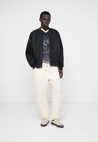 Black bomber jacket over a dark mesh top, paired with loose white pants and beige sneakers. Minimalist design with neutral colors.