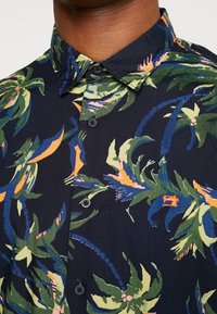 Mørkeblå kortærmet skjorte med et livligt tropisk bladmønster i grøn, orange og pink, med sorte knapper og spidse krave.