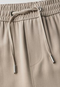 Pantalons de jogging beige avec une taille élastique, fermeture à cordon de serrage et embouts métalliques ; comporte une poche latérale et un tissu lisse et texturé.