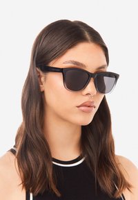 Musta suorakaiteen muotoinen aurinkolasit, joissa on tummat linssit ja paksu kehys. Brändin logo "HAWKERS" sangan kohdalla. Sileä, kiillotettu pinta, moderni muotoilu.