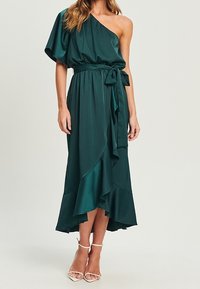 Vestido verde asimétrico de un solo hombro con cinturilla ceñida, falda fluida y dobladillo con volantes. Material de satén suave con un detalle de cinturón.