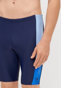 Morske kratke hlače za plavanje z svetlo modrim stranskim panelom, ki vsebuje geometrijski vzorec in logo Speedo. Narejene so iz gladkega, raztegljivega materiala.
