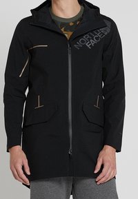 Giacca esterna nera con zip frontale, tasche laterali e cappuccio. Presenta dettagli a contrasto in marrone e un logo sul petto.