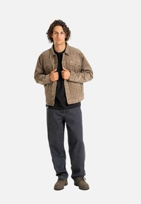 Chaqueta de pana marrón con un corte relajado, cierre de botones en la parte delantera y dos bolsillos en el pecho, combinada con una camisa negra y jeans oscuros. Zapatos marrones de cordones.