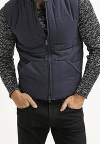 Marinblå vadderad puffer-väst med dragkedja fram och två sidofickor, lagd över en mönstrad stickad tröja, ihop med svarta jeans.