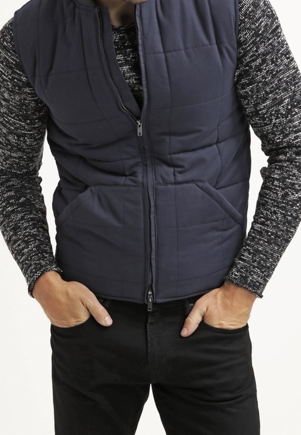 Námořnická prošívaná péřová vesta s předním zipem a dvěma bočními kapsami, vrstvená přes vzorovanou pletenou košili, v kombinaci s černými džíny.