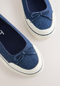 Paire de chaussures en denim bleu à enfiler avec semelles en caoutchouc blanc et bouts renforcés, chacune ornée d'un petit nœud sur le dessus.