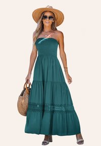 Turquoise, strapless maxi jurk met smock-buste, gelaagde rok en kanten randdetails. Gecombineerd met een gehaakt tas en een brede strooien hoed.