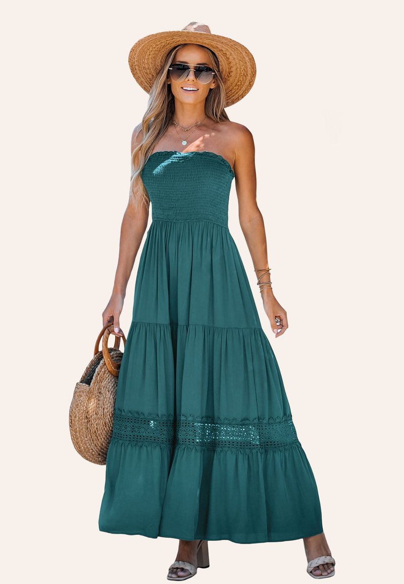 Turquoise, strapless maxi jurk met smock-buste, gelaagde rok en kanten randdetails. Gecombineerd met een gehaakt tas en een brede strooien hoed.