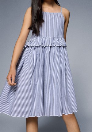 Mädchen trägt ein ärmelloses blau-weiß gestreiftes Kleid mit Rüschendetail und gewelltem Saum, steht vor einem grauen Hintergrund.