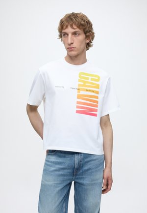 Νέος άνδρας που φοράει λευκό t-shirt Calvin Klein με κείμενο σε διαβάθμιση από κίτρινο σε κόκκινο και μπλε τζιν, στέκεται μπροστά σε απλό λευκό φόντο.