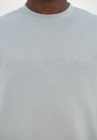 Gros plan d'une personne portant un t-shirt gris clair avec un texte en relief "ARMANI EXCHANGE" sur la poitrine.