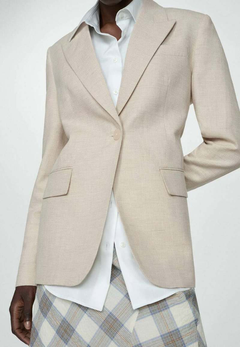 Beige, strukturiertes Blazer mit Revers, Ein-Knopf-Verschluss, seitlichen Taschen, kombiniert mit einem weißen Hemd und einem karierten Rock.