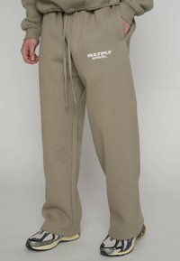 Beige Jogginghose mit "MULTIPLY APPAREL" Logo am Oberschenkel, kombiniert mit mehrfarbigen Sneakern, Hand der Person in der Tasche vor schlichtem Hintergrund.