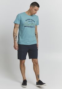 Lyseblå bomulls t-skjorte med grafisk "Blend Denim Surfing" trykk, kombinert med mørk marineblå cargo-shortser og svarte sneakers.
